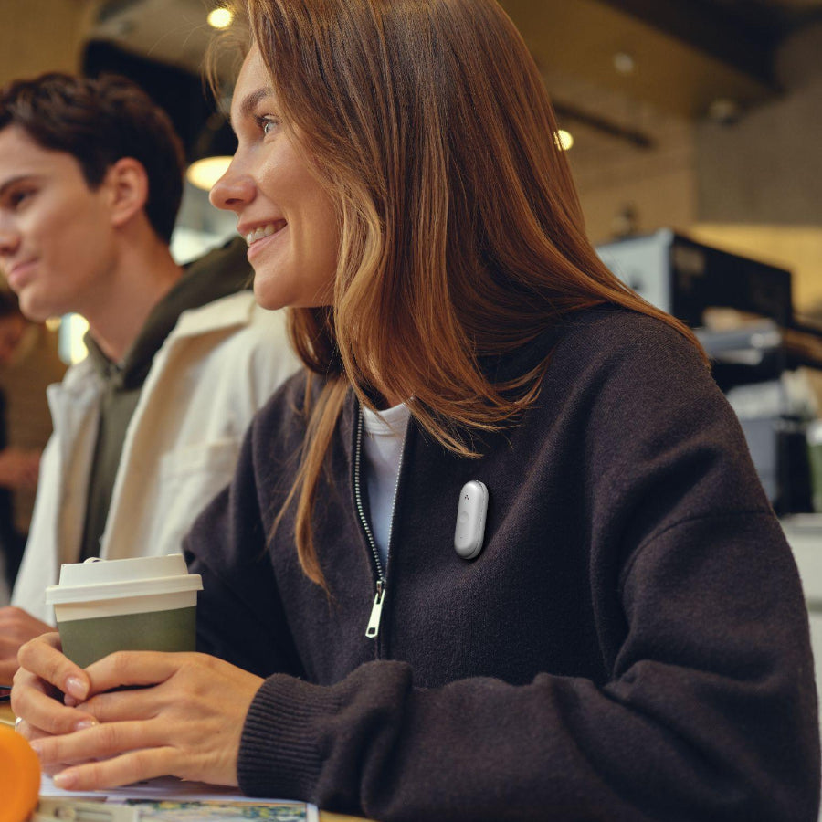 A woman in a cafe wearing a silver Plaud NotePin S on her dark jacket while holding a coffee cup.