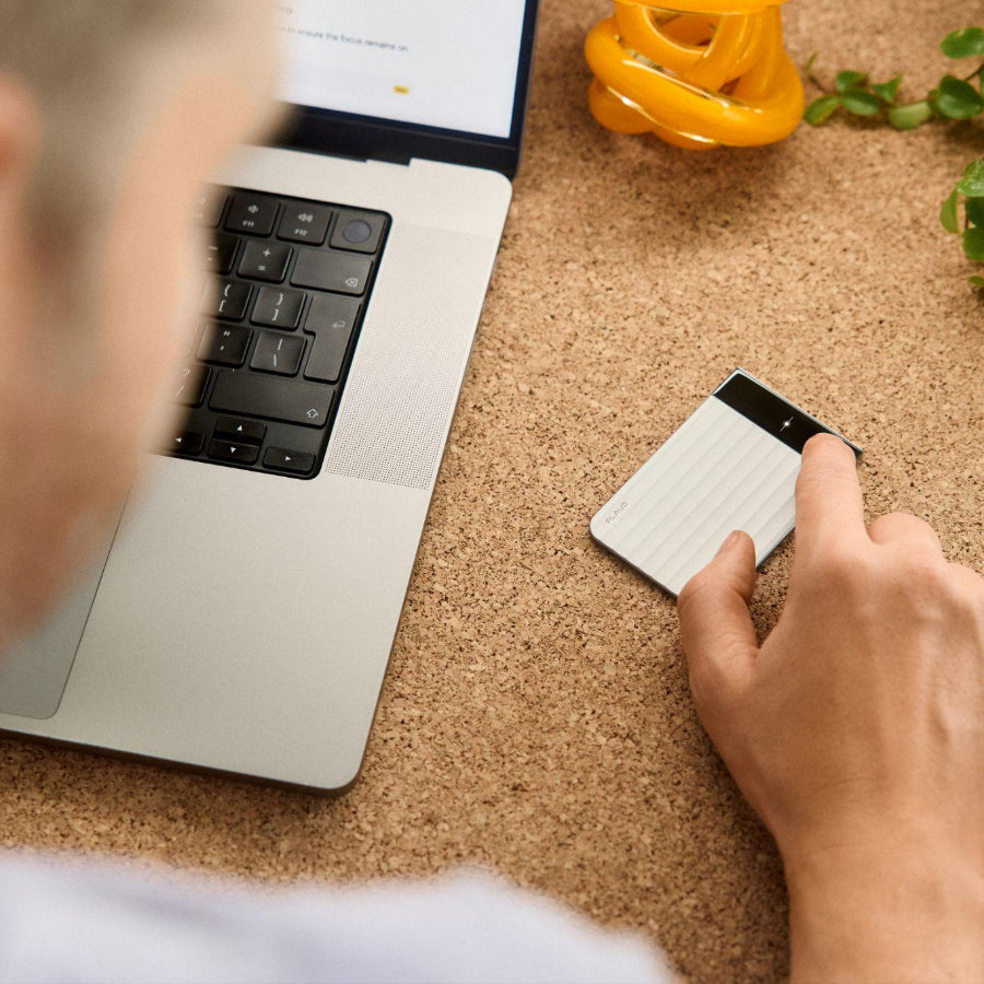A person using a silver Plaud Note Pro with a ribbed body and display screen on a cork desk next to a laptop.