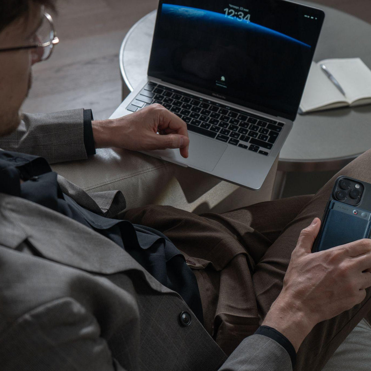A man in a suit holds a smartphone with a black Plaud Note attached while working on a laptop.