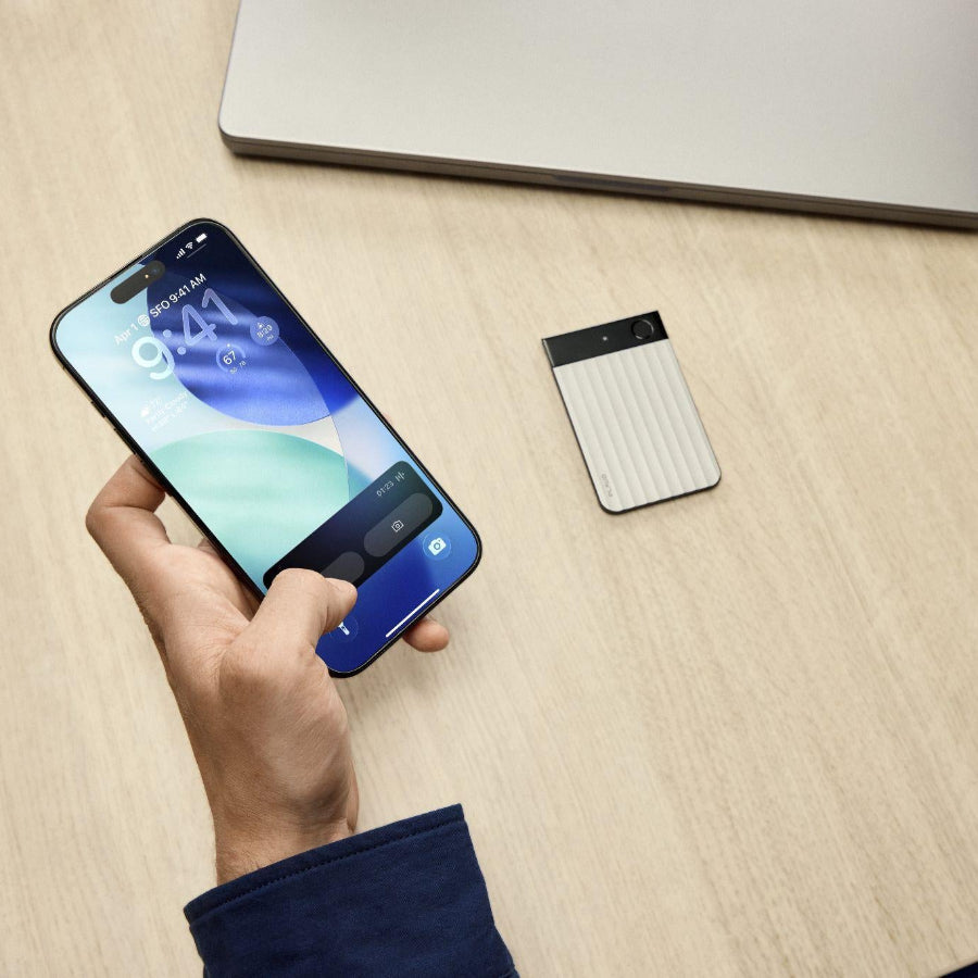 A silver Plaud Note Pro with a ribbed texture on a desk next to a hand holding a smartphone.