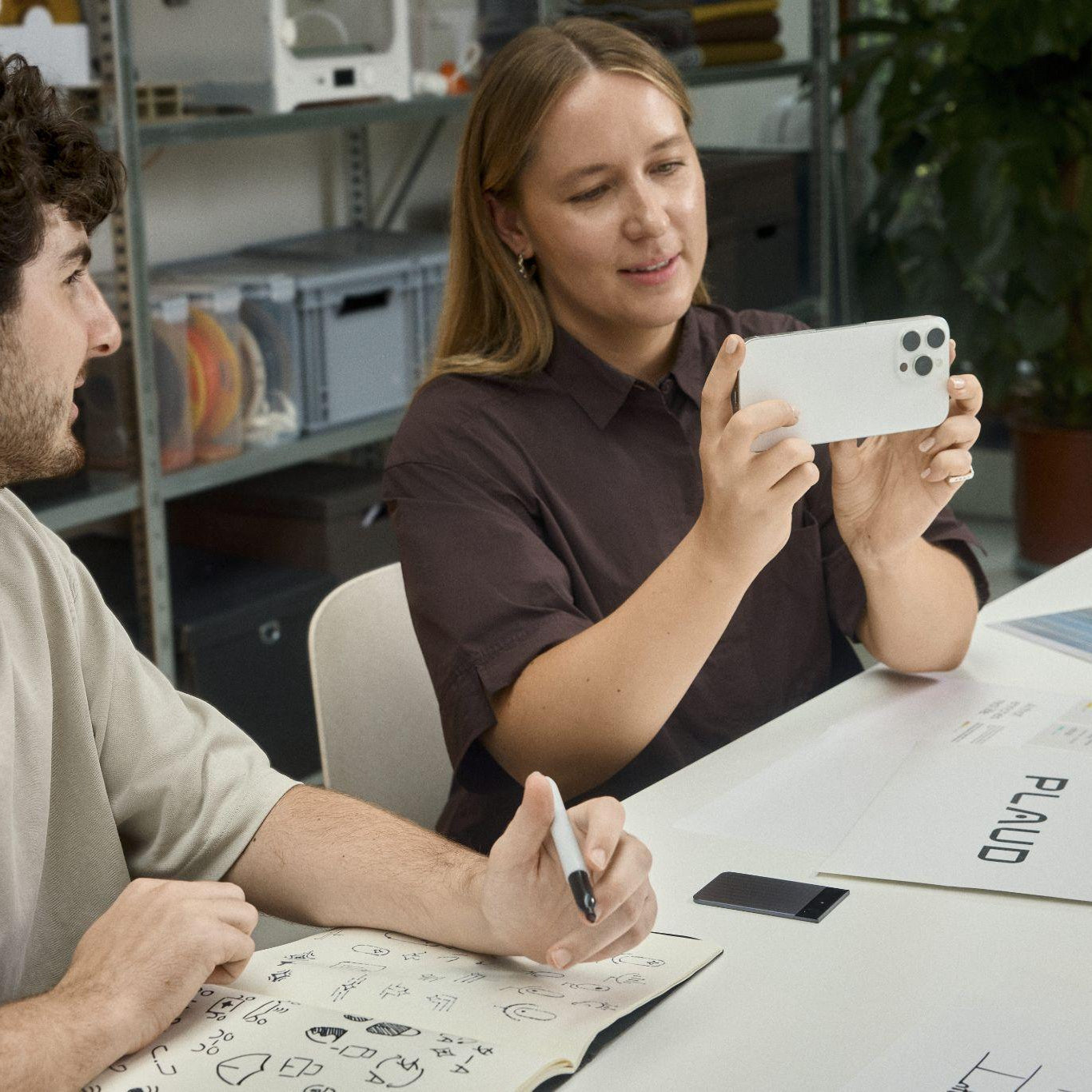 Woman photographing sketches next to a black Plaud Note Pro recording a meeting on a table.