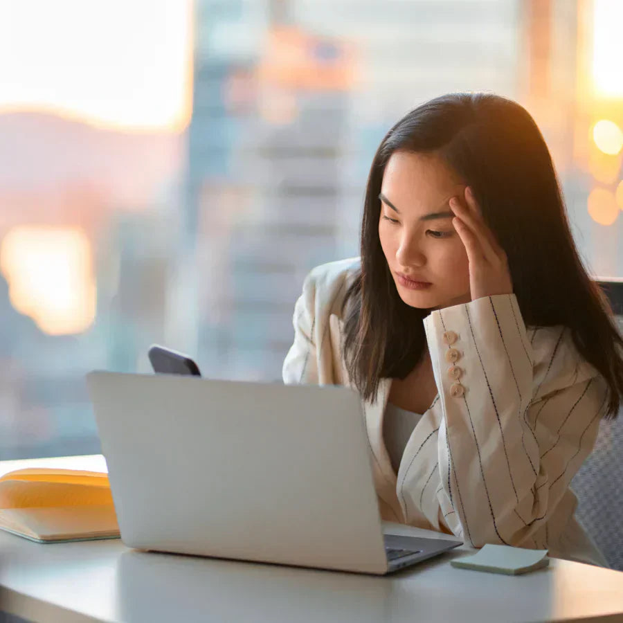 Stressed woman in a striped blazer looking at a laptop in a bright office.