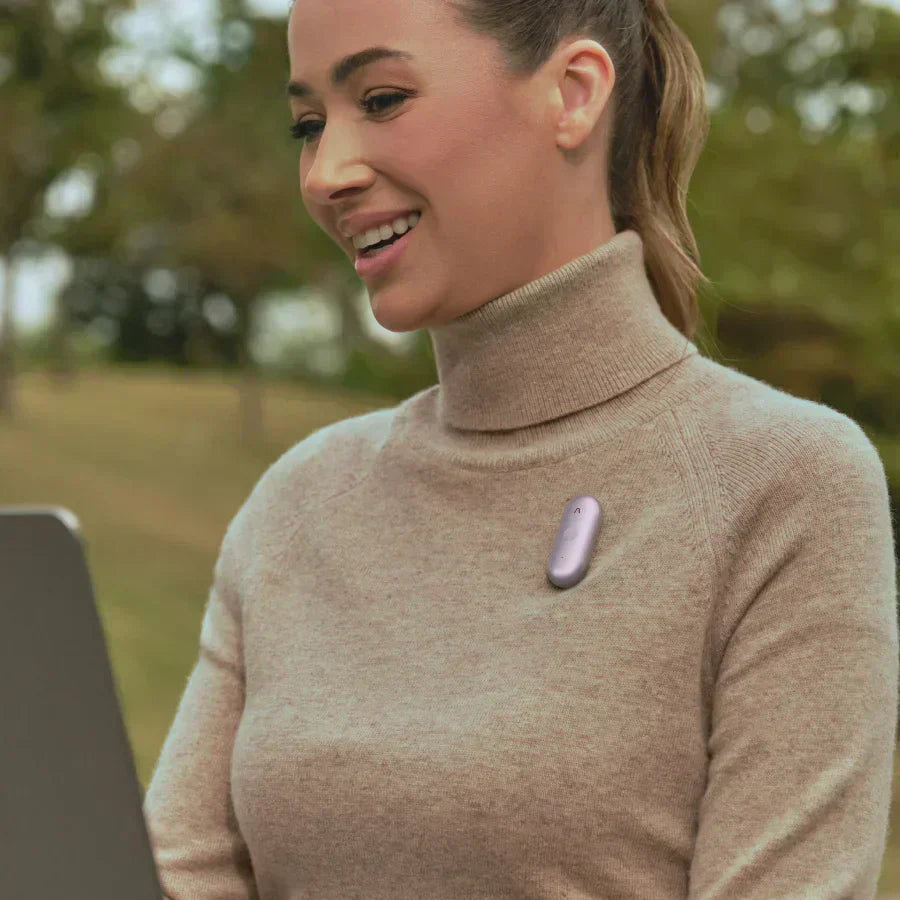 Woman wearing a purple Plaud NotePin S on her turtleneck sweater while using a laptop outdoors.