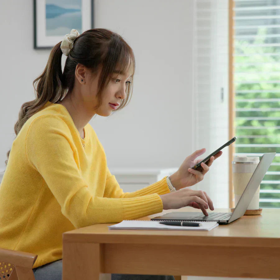 Woman in a yellow sweater using a laptop and holding a phone with a black Plaud Note attached to the back.