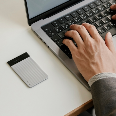 Silver Plaud Note Pro with ribbed texture and screen on a desk next to a person typing on a laptop.