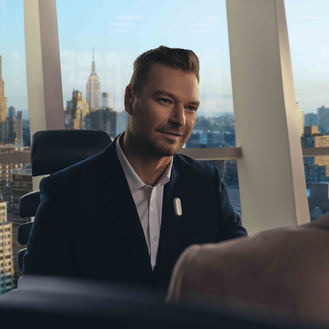 A man wearing a silver Plaud NotePin on his suit lapel in a high-rise office with a city view.