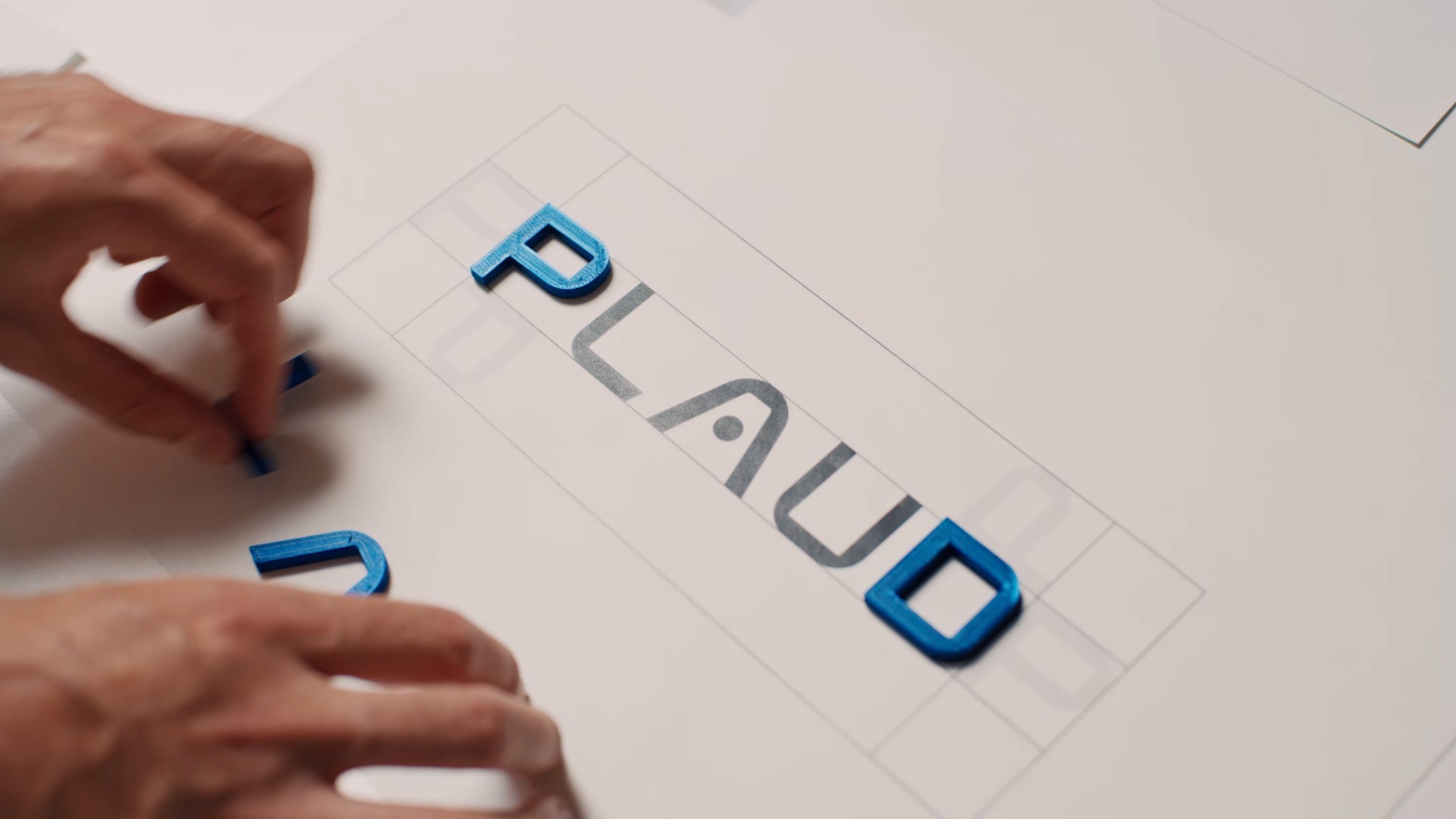 Hands arranging blue 3D letters to spell the Plaud brand name on a white design grid.
