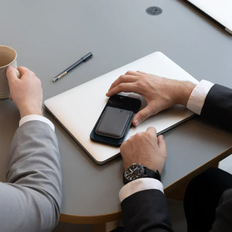 A professional holds a smartphone with a black Plaud Note attached, sitting at a desk with a laptop.