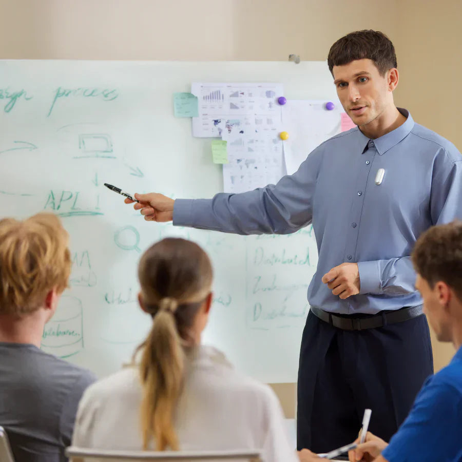 A presenter wearing a silver Plaud NotePin on his shirt during a meeting in front of a whiteboard.