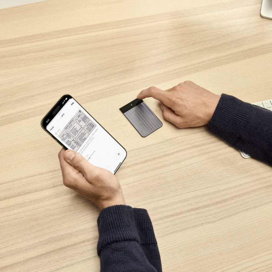 A person using a black Plaud Note Pro on a wooden desk while viewing meeting notes on the Plaud App on a smartphone.