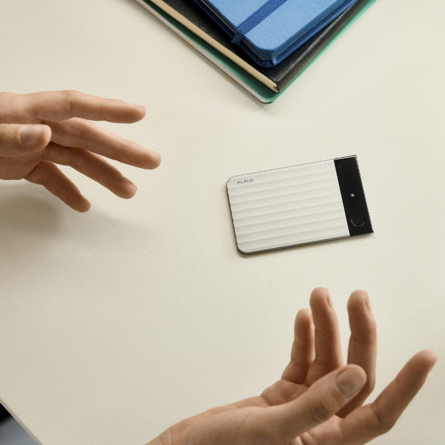 Silver Plaud Note Pro AI voice recorder on a desk between gesturing hands during a meeting.