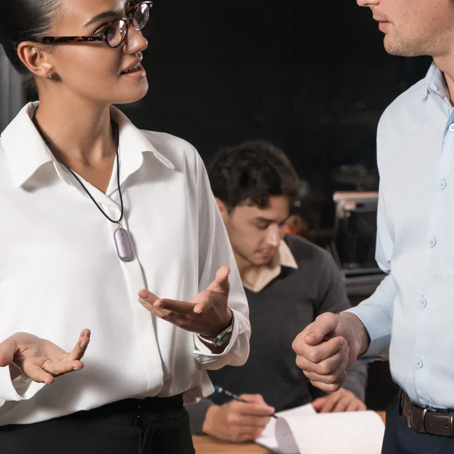 A woman wearing a purple Plaud NotePin S  on a lanyard while speaking with a colleague in a professional setting.