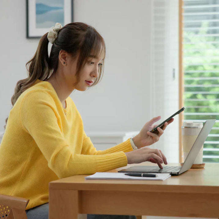 Woman in a yellow sweater using a laptop and holding a phone with a black Plaud Note attached to the back.