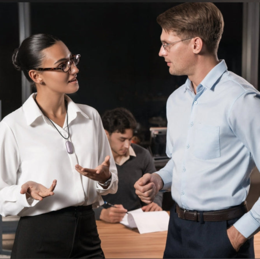 A woman wearing a purple Plaud NotePin on a lanyard talks to a colleague in a professional office setting.