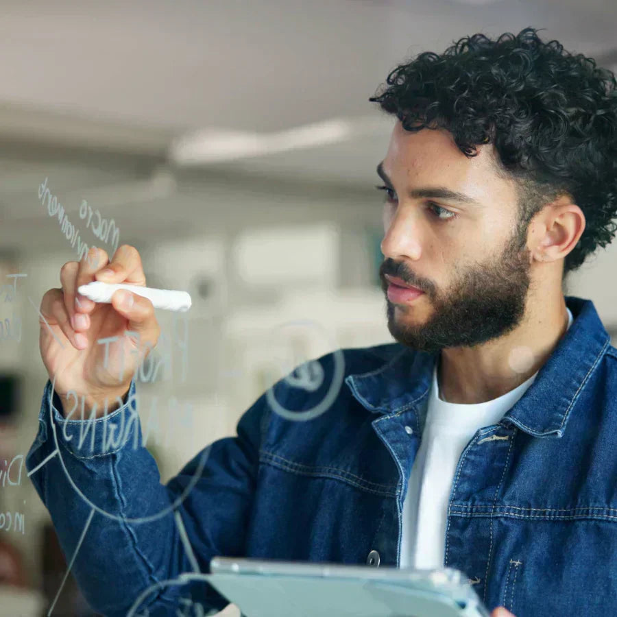 A man wearing a silver Plaud NotePin on his collar while writing on a glass board during a brainstorming session.