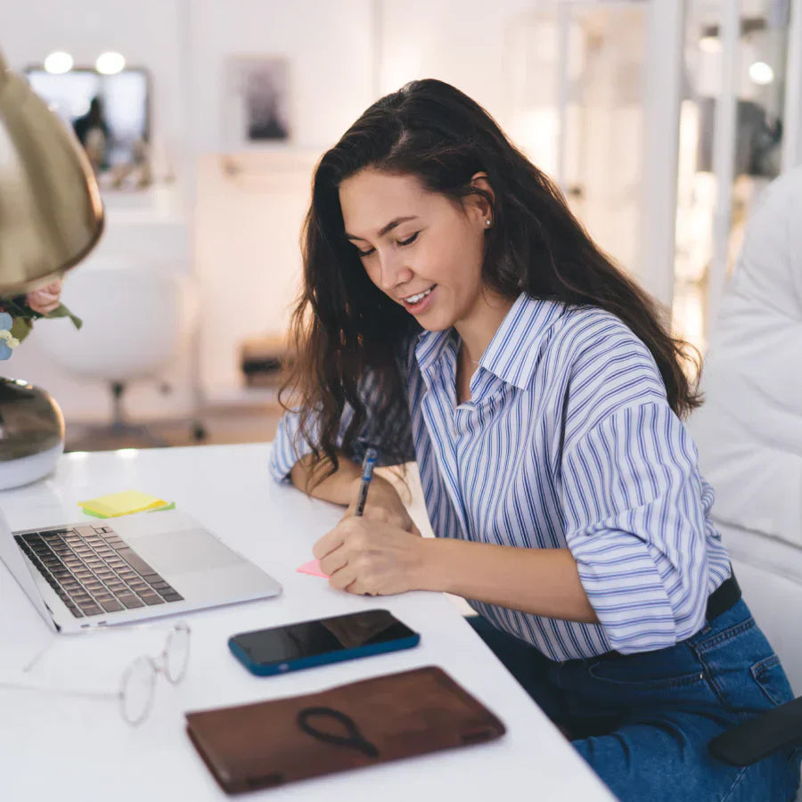 A woman writing at a desk next to a smartphone with a black Plaud Note Pro AI voice recorder attached.