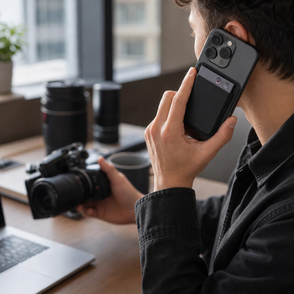 A person using a smartphone while holding a camera and a cup on a desk.