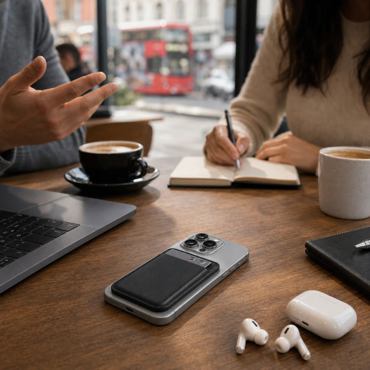 A smartphone, coffee cups, and a laptop on a wooden table with two people engaged in discussion.