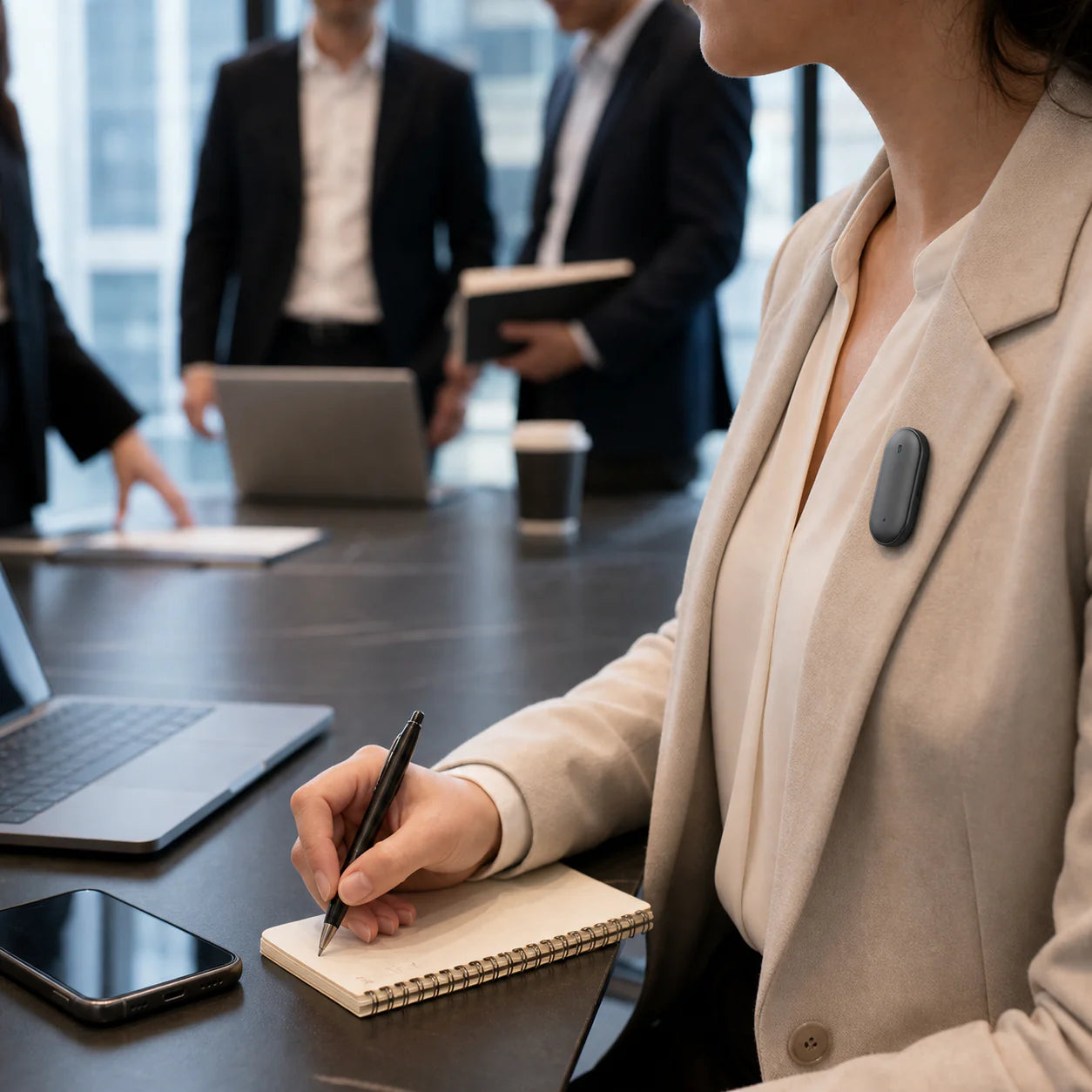 A person writing in a notebook while wearing a wearable device on their clothing, with a group of people in business attire in the background.