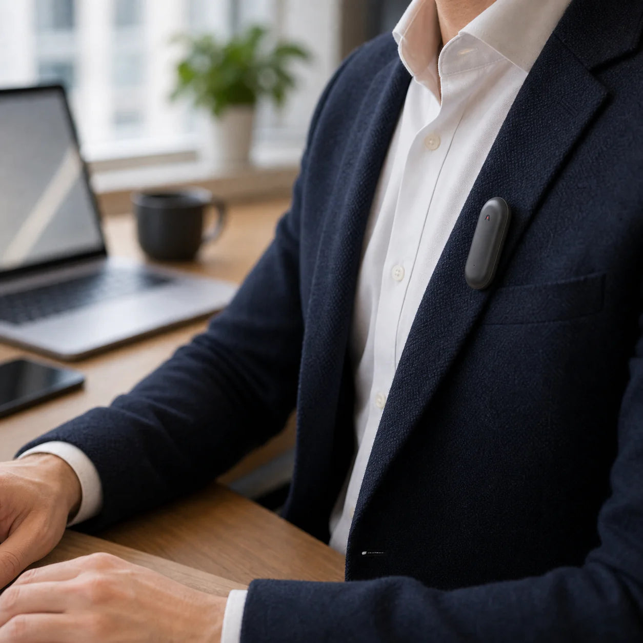 A person in a navy blazer with a white shirt sitting at a desk with a laptop, a phone, and a coffee cup. A wearable device is clipped to their shirt.