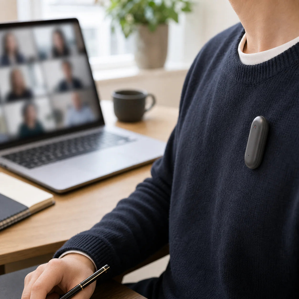 A person wearing a wearable device on their sweater while sitting at a desk with a laptop and notebook.