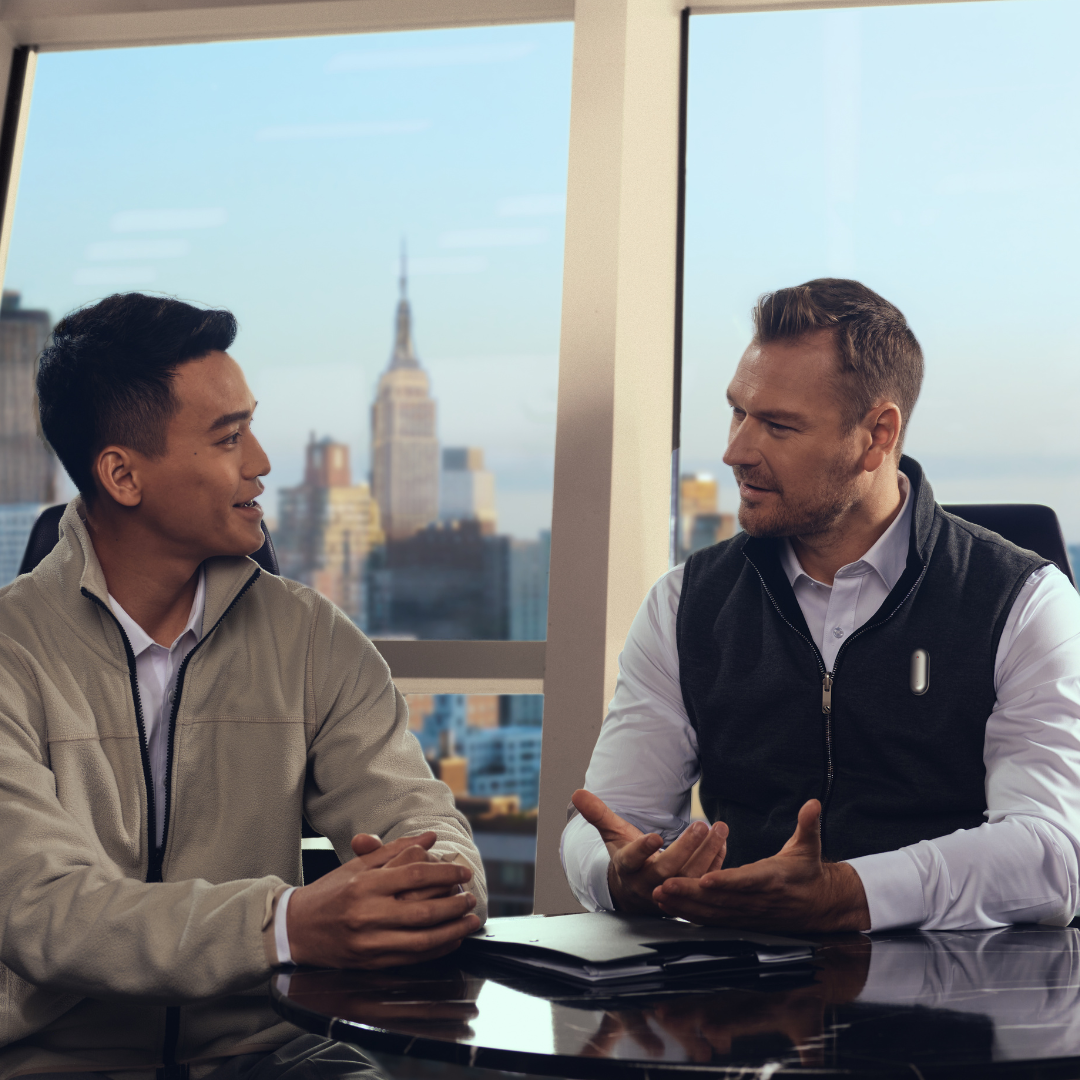 Two men in a business meeting with a city view. A silver Plaud NotePin is worn on a vest to record the conversation.