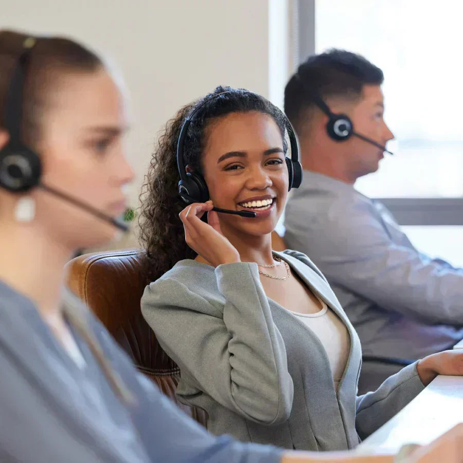 A smiling customer support representative wearing a headset in a busy office environment.