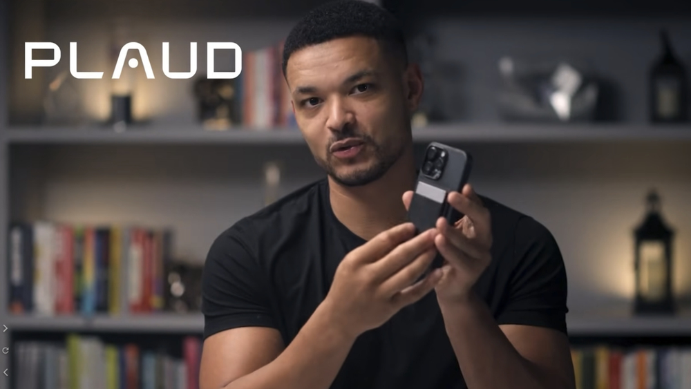 A man holds a smartphone with a black Plaud Note attached to the back in a home office setting.