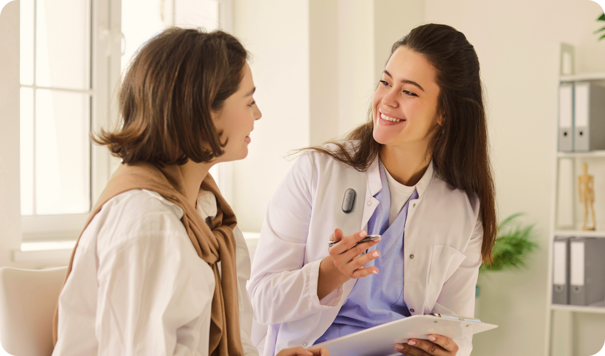 A smiling doctor in a white coat and blue scrubs speaks with a patient while wearing a Plaud wearable AI recorder on her lapel.