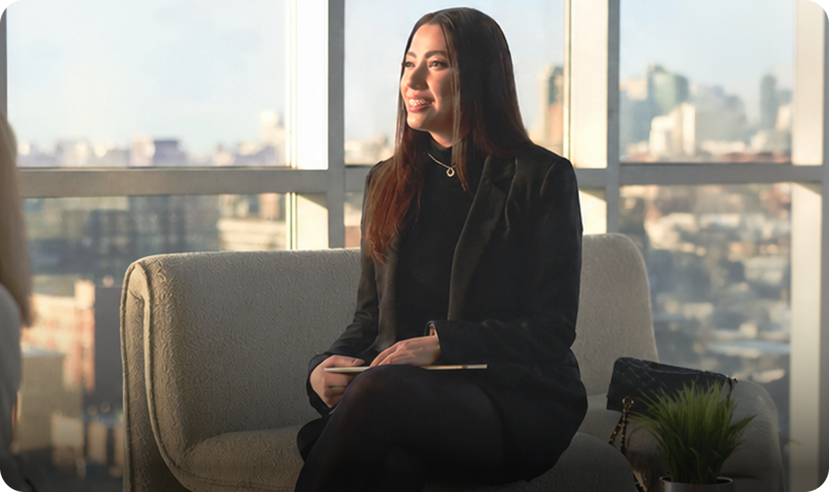A professional woman in a black blazer sits on a couch in a sunlit office with a city view, holding a Plaud AI voice recorder.