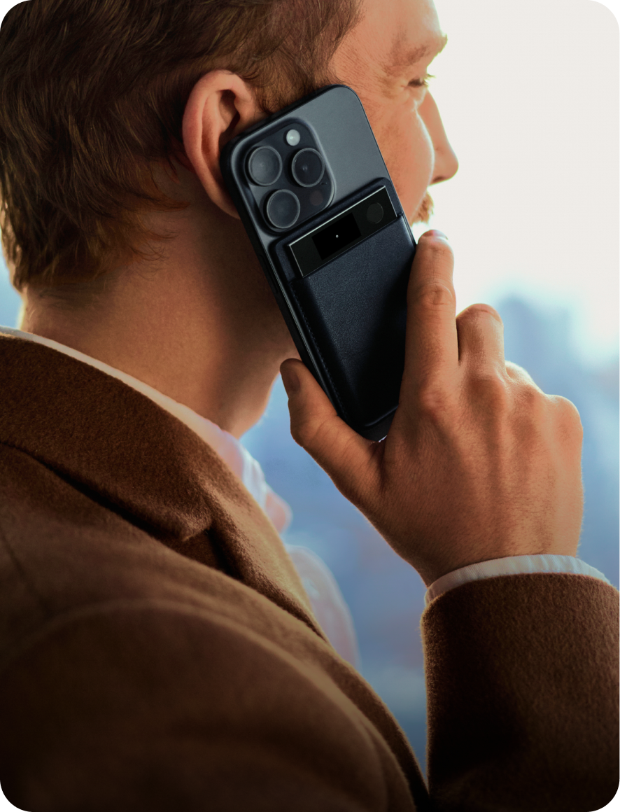 A man in a brown jacket holds a black smartphone to his ear with a black Plaud Note Pro attached to the back to record his call.