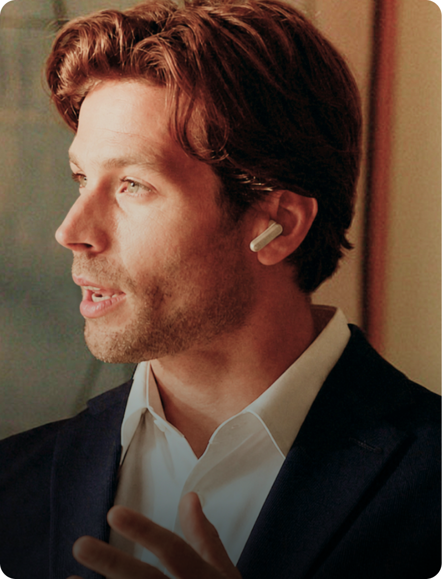 A man in a dark blazer gestures while speaking, wearing a white Plaud One earbud with a silver strip on the outer stem.