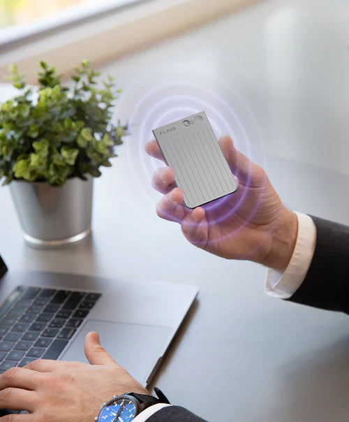 A hand holding a silver Plaud Note with purple signal waves over a laptop and desk.