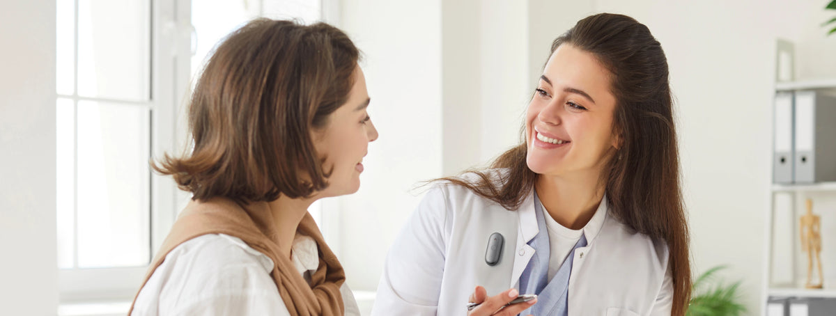 A doctor wearing a black Plaud NotePin S on her white coat smiles while speaking with a patient in a bright medical office.