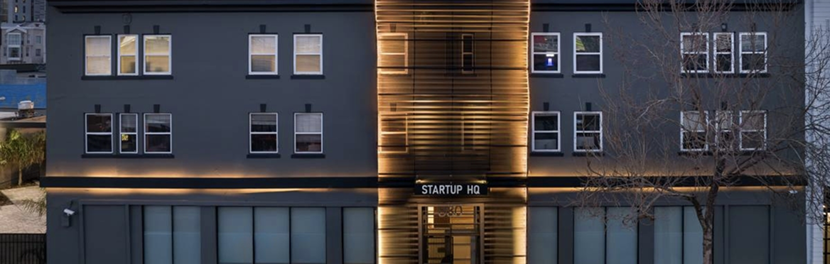 Modern dark grey office building at dusk with warm lighting and a sign reading STARTUP HQ.
