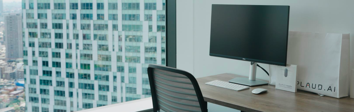 Modern office desk with a computer monitor, a Plaud.ai shopping bag, and a white Plaud NotePin box by a window.