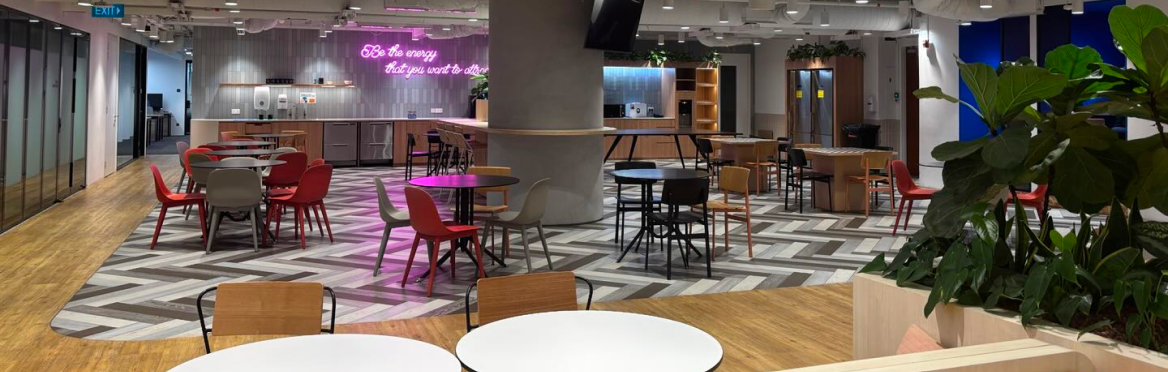 Modern office breakroom featuring colorful chairs, herringbone flooring, and a neon wall sign.
