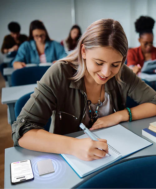 Student writing in a notebook with a silver Plaud Note and the Plaud App on the desk during a lecture.