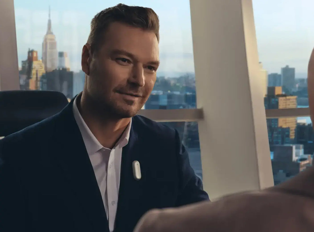 A man in a suit wearing a silver Plaud NotePin on his lapel during a business meeting in a high-rise office.