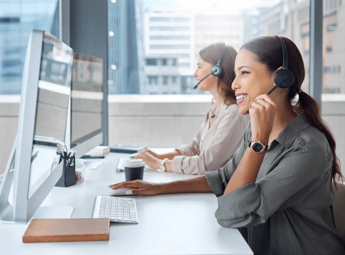 Two smiling customer support representatives wearing headsets while working at their computers in a bright office.